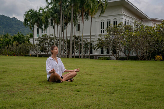Yoga of young woman meditating outdoors on the lawn under big tropical palms and beautiful house on background. Outdoor Classes takes place in the beautiful lawn