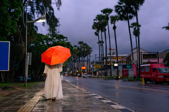 Back view of girl in white dress holding orange umbrella walking on sidewalk along road in Phuket