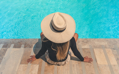 A woman wearing a straw hat sits on a stone staircase by a pool