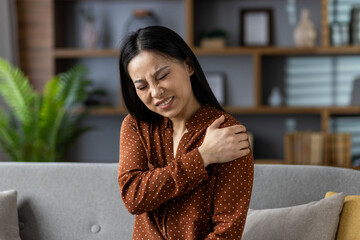Asian woman sitting on sofa with shoulder pain, seeking relief. Expression shows discomfort,...