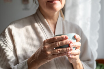 Mature caucasian woman is drinking tea or coffee from cup near window at home. Concept of domestic lifestyle. Cropped photo of modern happy middle aged lady wearing bathrobe at sunny day.