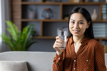 Asian woman sitting on sofa holding glass of water, smiling in cozy living room. Natural light and plant create relaxing atmosphere. Perfect for concepts of health, hydration, comfort.