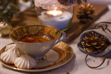 Winter still life. Cup of tea with meringues on a cozy table with a linen tablecloth on it. Beside a candle and decorative pine cones in a bag. Cozy atmosphere. Porcelain cup with gilding and painting
