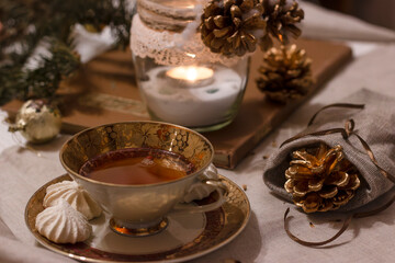 Cozy winter evening tea setting with cookies, pinecones, and candlelight at home. Cup of tea with cookies . Decorative pine cones on the linen tablecloth. Candle enhances the cozy atmosphere.