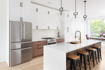 A kitchen with white and wood cabinets, bar stools sitting at a large island with a white marble waterfall countertop, stainless steel appliances, and white oak flooring. No brands or labels.