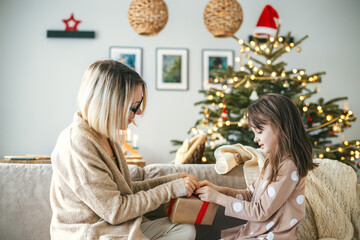 In a festive living room, a little girl and her mother sitting on a couch, opening a Christmas gift