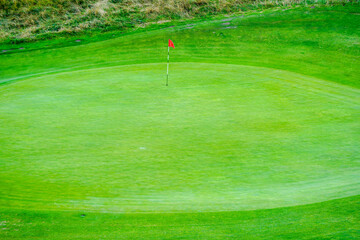 A pristine green golf course with a red flag marking the hole stands against a backdrop of lush grass