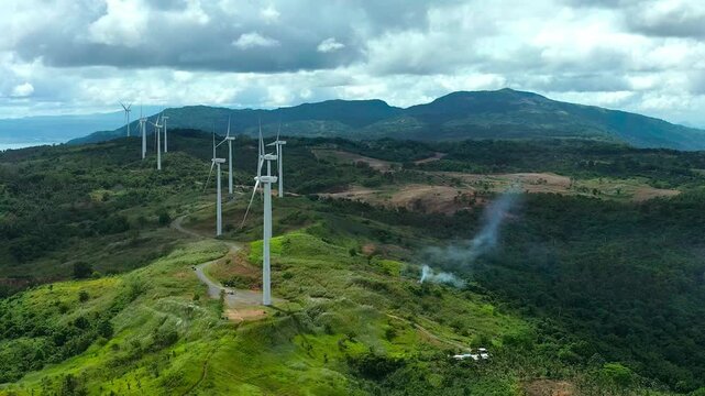 Pililla wind turbine farm on mountain during cloudy day. Burning wildfire in Philippines, Rizal. Aerial Wide shot.