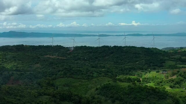 Aerial wide shot of green Asian island in Philippines in Rizal Pililla and rotating wind turbines in front of sea. Cloudy day in Asia. Aerial lateral wide shot.