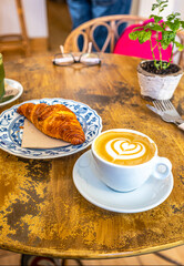 A Caffe Latte art and a croissant, served on a rustic wooden table, accompanied by a potted plant in the background.