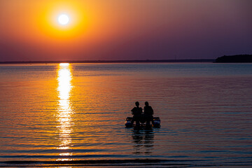 couple of young people are sitting on catamaran on background of multicolored sunset sky, sun disk and sunset track on the water