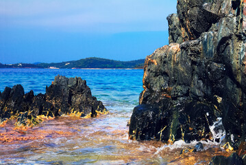 Beautiful scenic view - bright blue water of Aegean Sea, black sharp rocks, distant beach and pine wood, small island near Toroni, Sithonia of Chalkidiki (Halkidiki) peninsula, Greece, South Europe