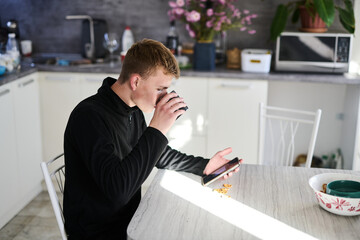 Obraz premium Young man sitting in the kitchen in the morning, drinking coffee and checking the news on his smartphone
