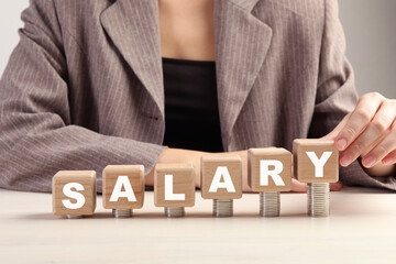 Woman putting wooden cubes with word Salary and stacks of coins on white table, closeup