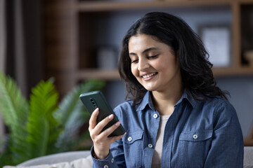 Close-up photo of a young Indian woman in a denim shirt at home, sitting on a sofa and using a mobile phone with a smile