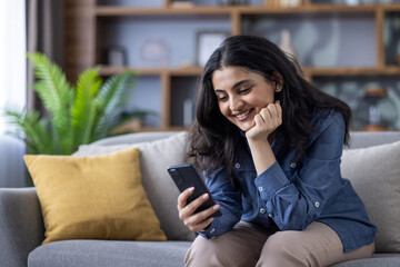 A dreamy and pensive young Indian girl is sitting on the sofa at home, resting her head on her hand...