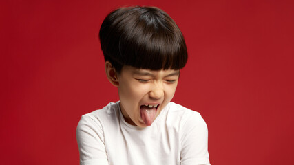 Little Asian boy closed his eyes and sticks out his tongue in silly, playful expression against...