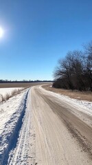 Fototapeta premium A long, winding road stretches through expansive snowfields, flanked by deep drifts. The bright sun casts a warm glow, creating a serene winter landscape.