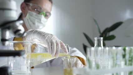 Woman scientist wearing a lab coat, white gloves, and a mask is pouring a yellow oily liquid from one beaker to another in a laboratory setting, close up. Science and medicine
