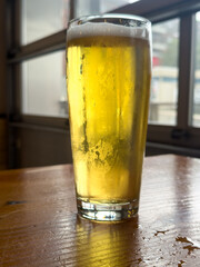 A tall clear pint beer glass filled with cold foam from a lager ale. The golden colored ale sits on the edge of a wooden patio table at a microbrewery. There's foam on top of the chilled glassware.