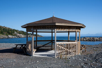 An open sided gazebo with a brown shingled roof, wood posts, and railings. The multi-sided structure or pergola overlooks the ocean with a deep blue sky. The park beach in the rocky background.