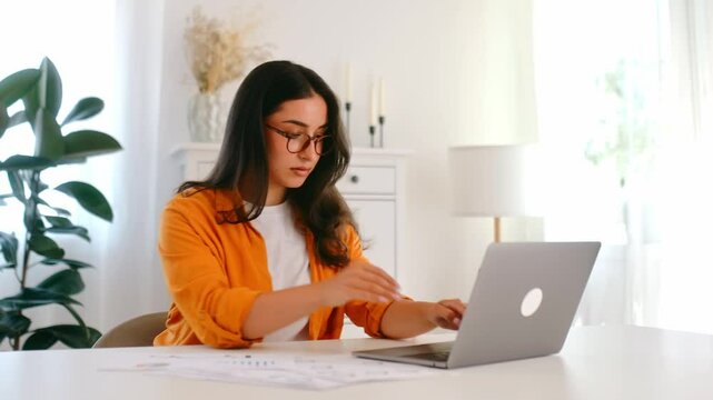 Focused successful pretty arabian or indian ethnicity young woman in an orange shirt, IT specialist, product manager sits at a workplace working in a laptop on a project, looking device screen, smiles