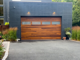 A modern brown faux wooden exterior garage door with four small horizontal glass windows. The modern door is on a luxury dark grey contemporary house with a concrete driveway. 