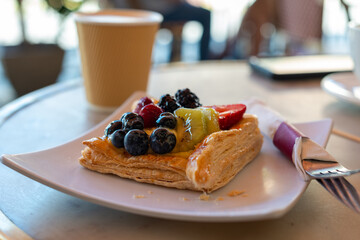 A paper cup filled with hot coffee on a round table with a white porcelain plate, fork, knife and napkin. On the plate is a square fruit pastry with blueberries, strawberries, blackberries and kiwi.