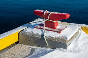 A colorful red steel mooring is wrapped with new teal green rope on a pier. There's snow on the wharf and there's water in the background.  The moorings are screwed into the wharf with large bolts.