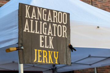 A large black outdoor sign with kangaroo, alligator, elk, and jerky in white and yellow text. The meat variety is for sale. The signage is on a pole next to a white tent at a farmer's market. 