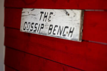 Fototapeta premium A white wooden sign with black letters indicates the gossip bench. The signage is attached to an exterior red wooden wall of a shed with white trim. The gossip bench is for meetings and networking.