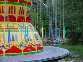 Empty old-fashioned merry-go-round in a city park. Red and blue wooden seats on metal chains of swing carousel. Trees on the background, fallen leaves on the ground © Olena