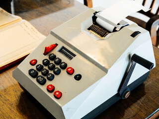 A vintage mechanical adding machine with a crank handle and numeric keypad sits on a wooden desk...