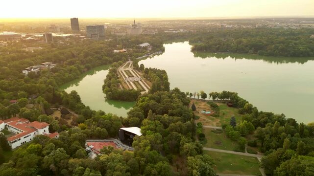 High angle drone footage of Herastrau and Arcul de Triumf in Bucharest Romania. Green park shot on a sunny day during golden hour. Cityscape of warm summer evening. Rugby stadium visible from above
