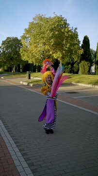 Street performer in a vibrant costume juggles colorful ribbons while walking gracefully through a sunny park in slow motion
