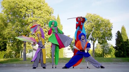 Group of street performers in colorful costumes juggle and pose on stilts in a park, captured in slow motion