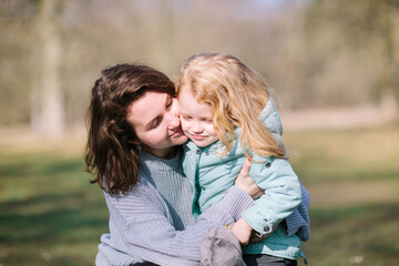 Fototapeta premium Young beautiful mother and an adorable blond little daughter are hugging and kiss each other in a park, happy family