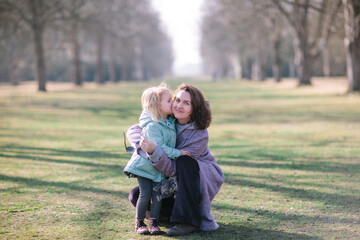 Young beautiful mother and an adorable blond little daughter are hugging and kiss each other in a park, happy family