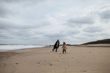 Mother in a warm coat and little daughter in a trench coat run after each other and have fun on the sandy beach with a strong wind in autumn, Norfolk, England