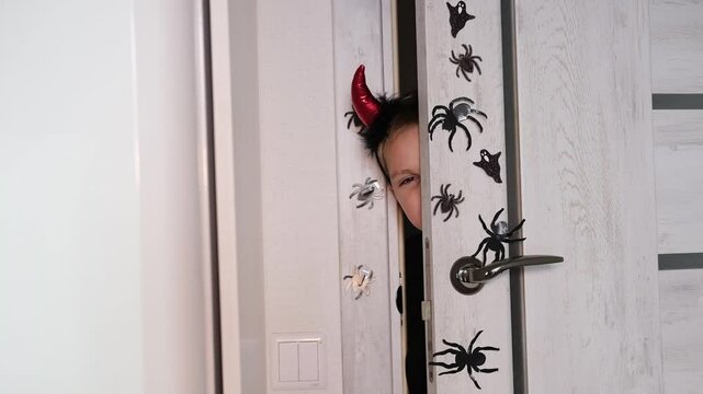 Beautiful little devil with horns. Baby. Concept of Halloween, holiday and childhood. Funny child in carnival costume looks out from behind door of house. Selective focus.	