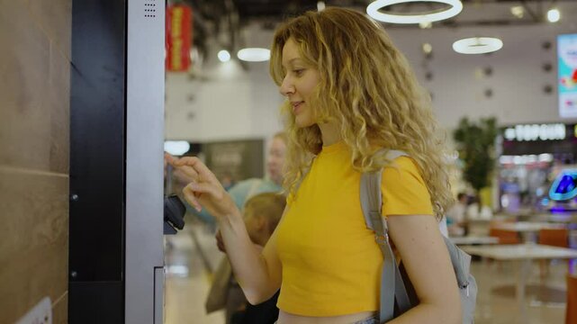 Young woman with curly blonde hair interacts with a digital self-service terminal, placing an order for food in a fast food restaurant, showcasing modern technology and convenience