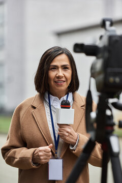 A multiracial female journalist holds a microphone, confidently reporting live from an urban location.