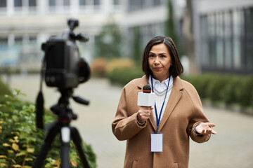 A multiracial female journalist holds a microphone, delivering news outdoors in a lively environment.