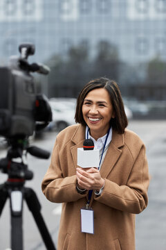 A multiracial journalist in a beige coat eagerly conducts an outdoor news report, microphone in hand.