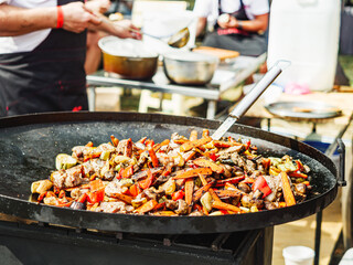 Street Food Festival: Man Stir-Frying Vegetables and Meat in a Large Pan at a Vibrant Urban Gathering