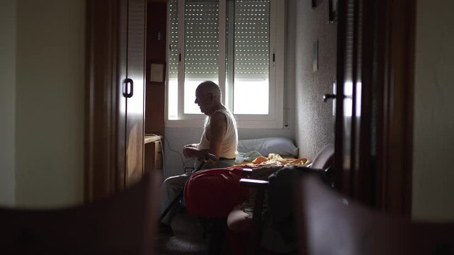 video of elderly man in his room in the morning, single bed and natural light. He gets up and walks with two walking sticks.