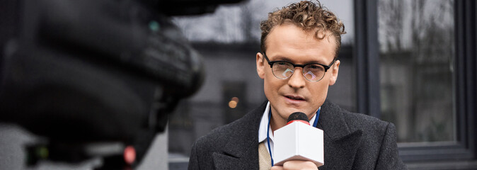 The journalist, wearing glasses and a coat, delivers insightful commentary into a microphone outdoors. © LIGHTFIELD STUDIOS