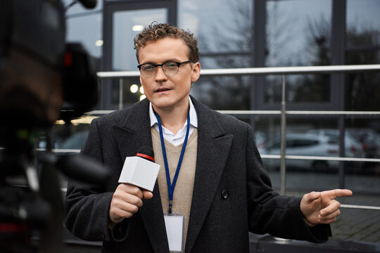 The journalist wearing glasses and a coat confidently speaks into a microphone, reporting news updates.