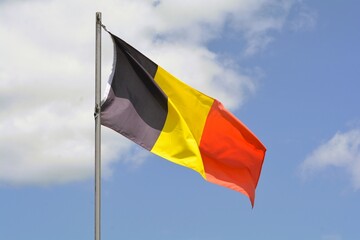 The national flag of Belgium flies up a flagpole on a beautiful summer day with a background of blue sky and white clouds.