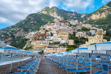 Naklejka premium Colorful buildings cascade down the hillside as visitors bask on Positano's sun-soaked shores, where iconic beach umbrellas dot the landscape beneath stunning cliffs. 